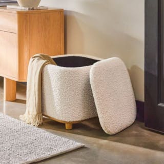 A cream bouclé storage ottoman with the lid open and a throw blanket draped over the side.