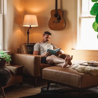 Tan leather armchair and ottoman in a cozy living room with warm lighting and a person reading a book.