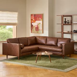 Chocolate brown leather corner sectional sofa with tufted seats and bolster pillows, styled with a glass coffee table and green rug.