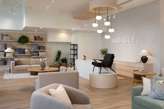 Modern showroom featuring a neutral-toned living room with sofas, armchairs, a coffee table, and wood storage furniture, set against a warm slatted wood feature wall with a TV and modern open shelving.