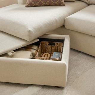 Beige reversible sleeper sectional with an open storage chaise holding blankets and a woven basket.