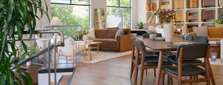 Modern dining area with a wooden table and black leather walnut chairs, surrounded by open shelving displaying nightstands in a warm, naturally lit showroom with big windows.