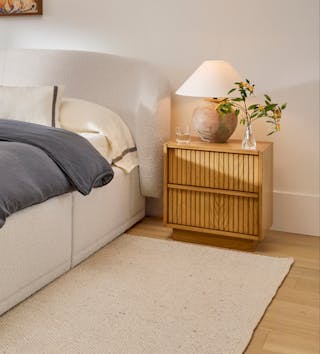 Kouva nightstand in natural oak with vertical wood detailing, beside a cozy upholstered bed.