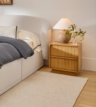 Kouva nightstand in natural oak with vertical wood detailing, beside a cozy upholstered bed.