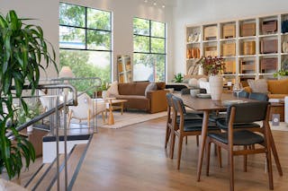 Modern dining area with a wooden table and black leather walnut chairs, surrounded by open shelving displaying nightstands in a warm, naturally lit showroom with big windows.