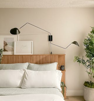 Cozy bedroom scene featuring a wooden slatted headboard with white and light grey bedding, framed art above the headboard, and a black wall-mounted arm lamp above.