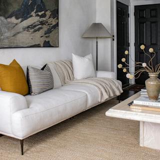 Quartz white Abisko sofa with tapered brushed aluminum legs, mustard and striped accent pillows, and a cream throw beneath framed mountain artwork in an organic modern living room.