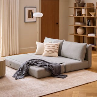 A light gray sofa bed with pillows and a throw blanket in a cozy living room.