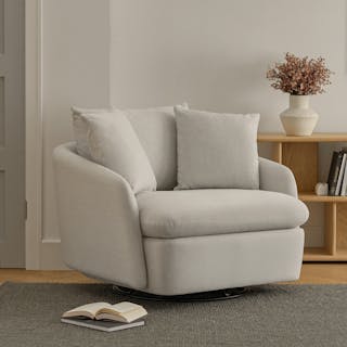 A silver ivory upholstered swivel chair with a curved silhouette and two plush pillows, styled in a living room. 