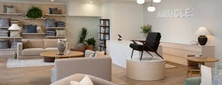 Modern showroom featuring a neutral-toned living room with sofas, armchairs, a coffee table, and wood storage furniture, set against a warm slatted wood feature wall with a TV and modern open shelving.