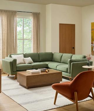 Timber corner sectional upholstered in Olio Green fabric with a wood base and cream accent pillows beside a rectangular wood coffee table in an organic modern living room.