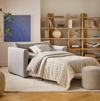 Light gray sleeper chair with a pull-out bed and striped bedding in a living room with wooden bookshelves.